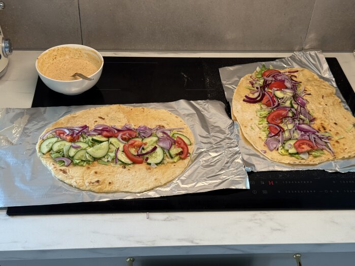 Hemgjorda kebabrullar med grönsaker och röd lök på bröd bredvid en skål sås på köksbänk.