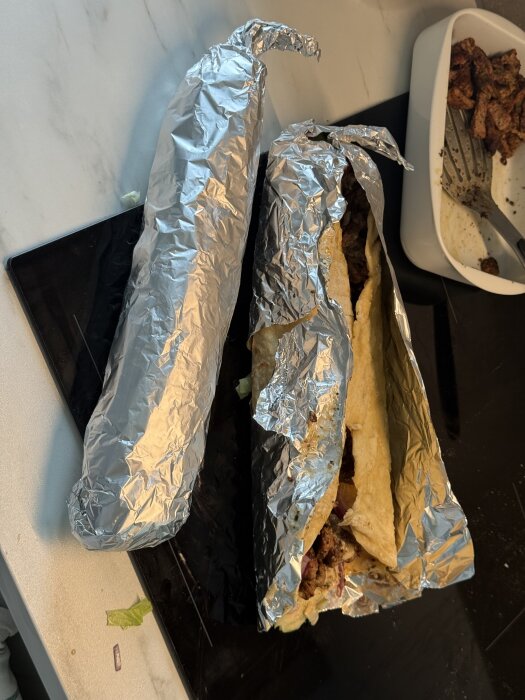 Hemgjorda kebabrullar inslagna i folie, med hemmagjord sås och bröd, ligger på en köksbänk bredvid en skål med kebabkött.