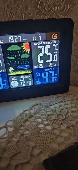 Digital väderstation visar inomhustemperatur 25°C, luftfuktighet 87%, klocka 19:27, under kylningsprocess. Bakgrund med mönstrad duk.