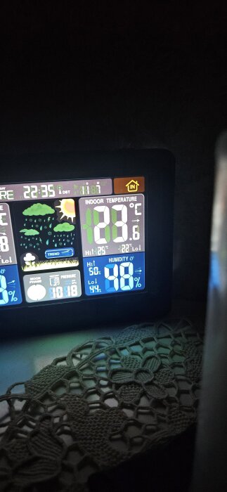 Digital väderstation visar inomhustemperatur 20,6°C och luftfuktighet 98%. Visar även datum och tid 22:35 samt vädersymboler och en virkad duk.
