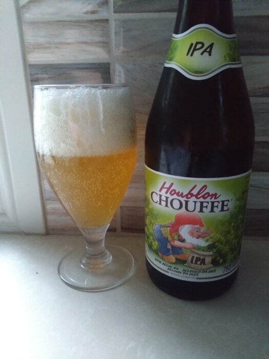 En flaska Houblon Chouffe IPA och fullt glas med skummande öl på ett bord framför kaklad vägg.