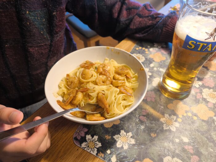 En person håller en tallrik med vitlöksräkor och pasta bredvid ett glas öl på ett blommönstrat underlägg.