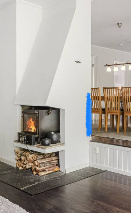 Öppen spis med vedförråd på vit vägg, blå markering visar var man överväger att borra för babygrind, matbord i bakgrunden, några trappsteg syns.