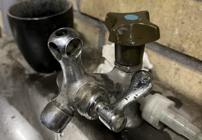 Gamalt, metallblandare med rost och kalkavlagringar i en snickeriverkstad, svart kopp i bakgrunden.