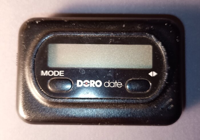 Svart personsökare med texten "Doro date" och två knappar på framsidan, ligger på ljus bakgrund.