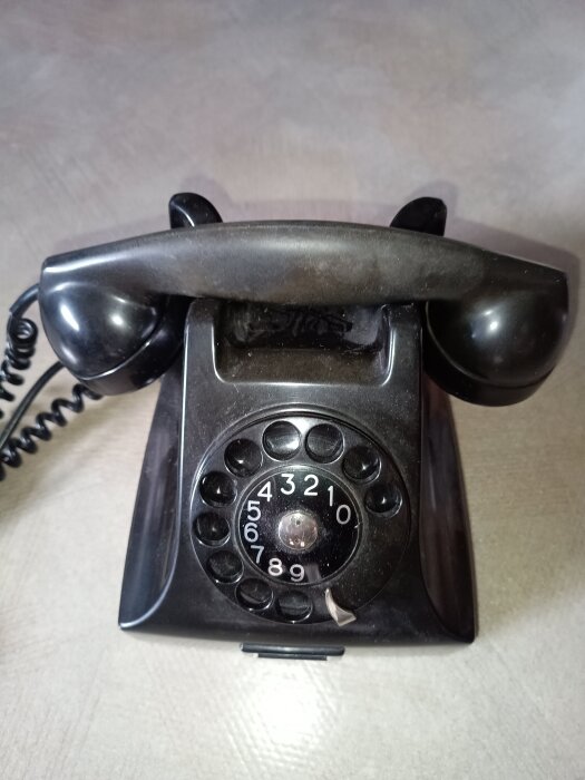 Svart gammaldags bakelittelefon med nummerskiva och lur, placerad på en bordsyta.