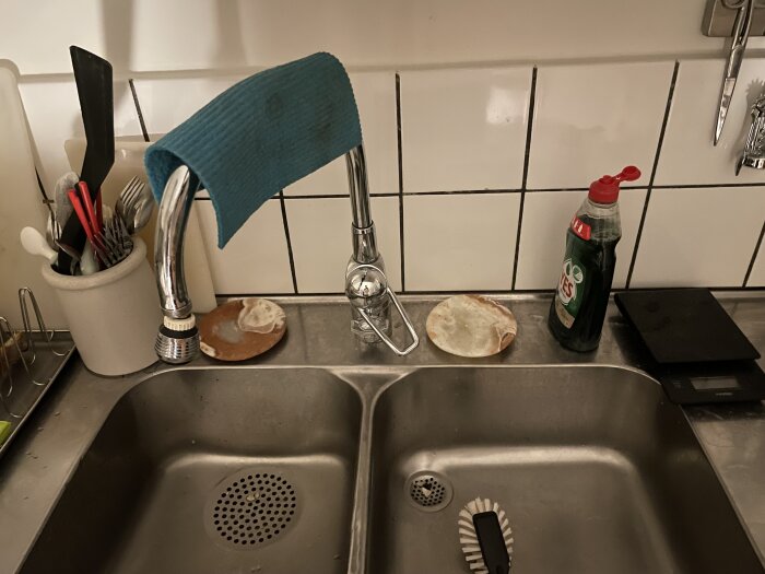 Diskbänk med två hoar, blandare med diskmaskinsavstängning och rengöringsmedel. Redskapskruka och diskborste synliga.