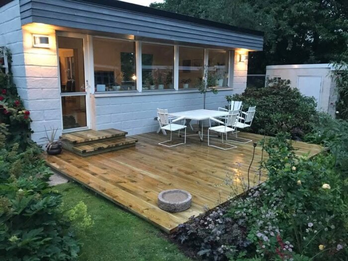 Trädäck vid hus med trallbrädor, uteplats med bord och stolar, omgiven av trädgård. Välbyggt med olika bredder på brädor.