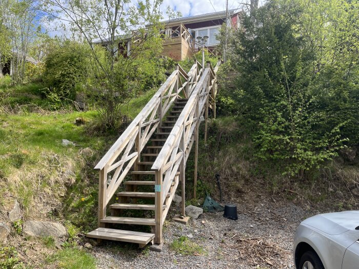 Trätrappa uppför en kulle, omgiven av grönska och träd, med ett hus i bakgrunden. Trappan är inte förankrad i marken längst ner.