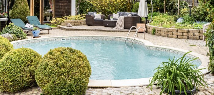Utomhuspool i en trädgård med omgivande växter, solstolar och utemöbler i bakgrunden.