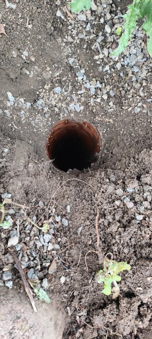 Brun dräneringsrör syns delvis under jord, omgivet av grus och jord, med grön vegetation vid kanten.