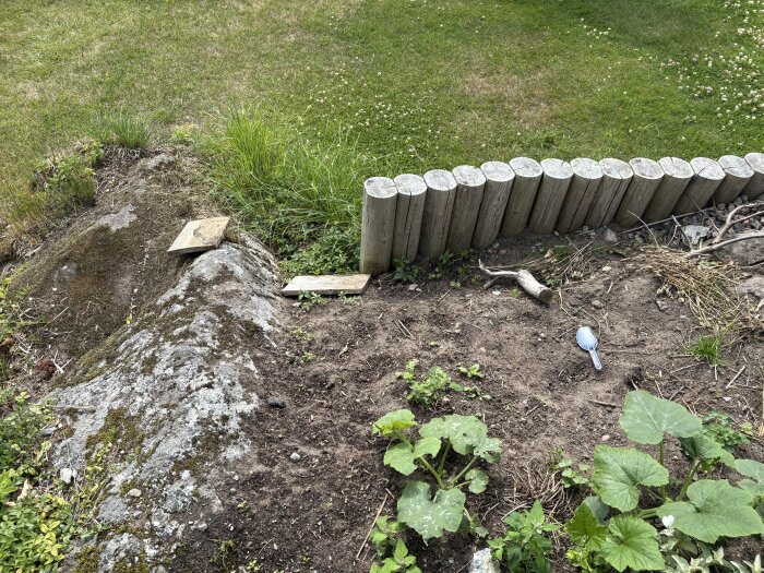 Trämur i trädgård med ojämnt berg och jord framför, träplankor och sked ligger på marken, gröna växter växer runtom.