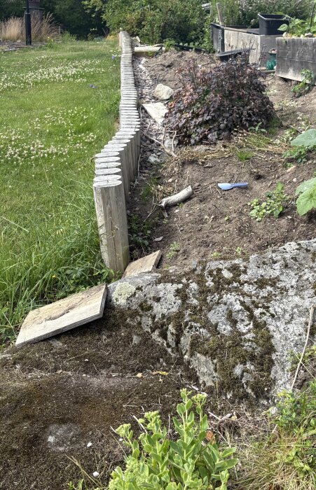 Trästockar bildar en lutande stödmur längs en gräsbevuxen sluttning med synliga stenar och en buske i bakgrunden.