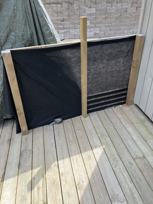 Byggprojekt med svart duk mellan trästolpar på trädäck för insynsskydd och vindgenomsläpp.