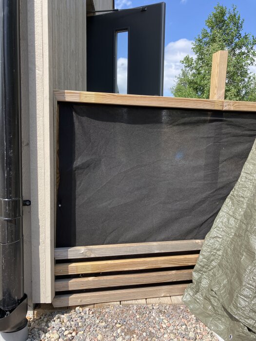 Träplank med svart insynsskyddsduk mellan ribbor, delvis uppställt bredvid ett hus med öppen dörr och grön presenning.