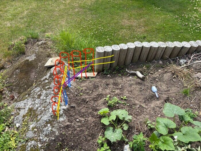 Anvisningar för att bygga en stödmur av timmer mot berg, med färgmarkeringar för montering och stabilisering, på en gräsmatta med makadam och växter.