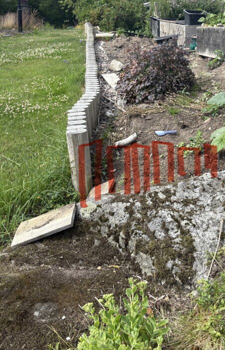 Trästockar bildar en låg trädgårdsmur mot en grässlänt och jord. En stenplatta och trädgårdsredskap syns på marken.