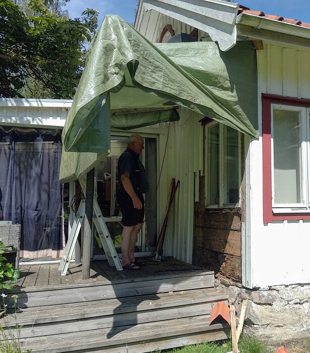 Man står på veranda under temporär presenning nära ett hus under renovering. Stege och verktyg synliga, väggparti saknas.
