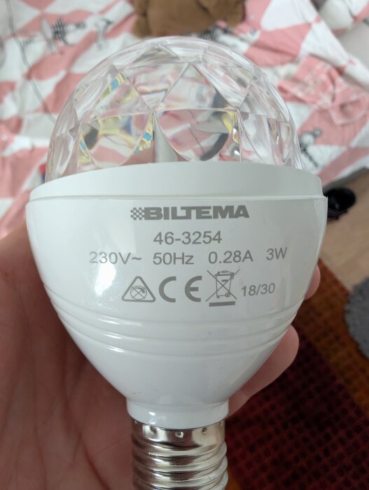 En dekorativ glödlampa från Biltema, som lyser i regnbågsfärger och roterar. Text visar specifikationer: 230V, 50Hz, 0.28A, 3W.