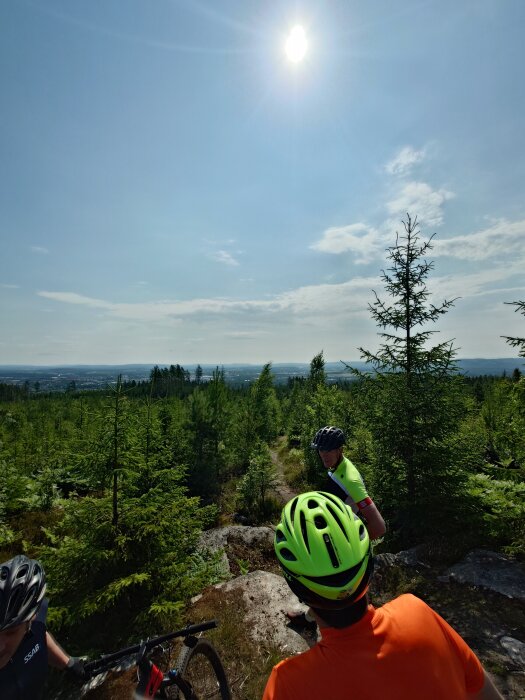 Cykelentusiaster på skogsstig, solig dag, utsikt över skog och stad i fjärran.