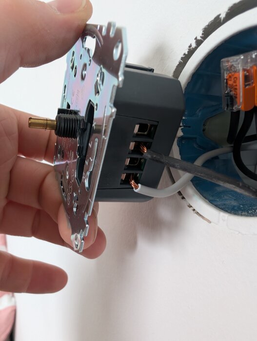 En person håller en lös elkontakt framför en infälld kopplingsdosa med en skruvmejsel som pekar mot exponerade koppartrådar.