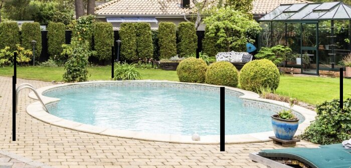 Rund pool i trädgård med planerade stolpar för att fästa ett nät runt. Vacker grönska, glasväxthus och stenlagd yta syns i bakgrunden.