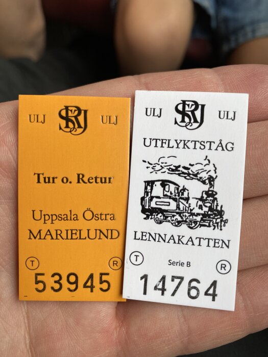 Två biljetter för ULJ-tågresa: en orange, "Tur o. Retur" Uppsala Östra - Marielund, och en vit, "Utflyktståg" Lännakatten, hålls i en hand.
