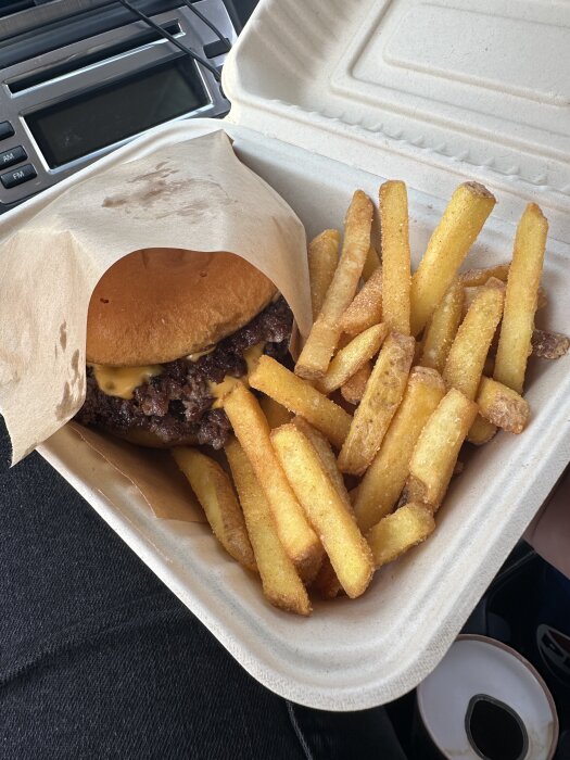 Hamburgare och pommes i en brun pappersförpackning, fotad inuti en bil.