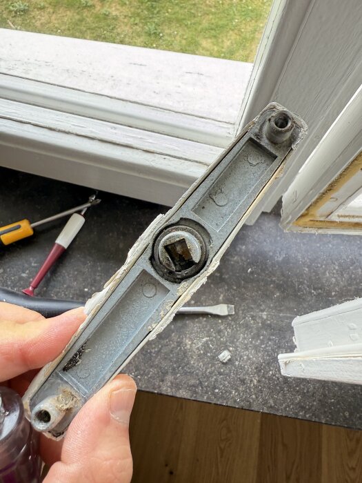 Närbild av ett löstaget fönsterlås med två stora skruvhål, placerat vid ett öppnat fönster med verktyg i bakgrunden.