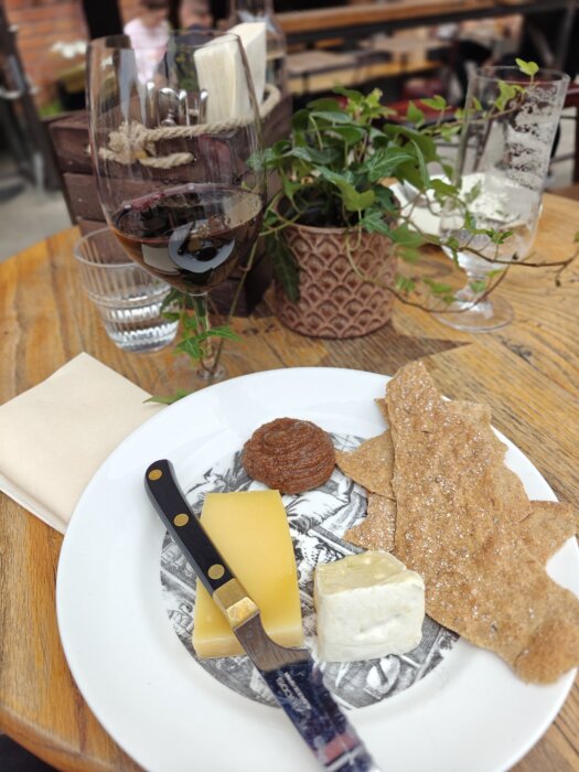Ostbricka med knäckebröd, fikonmarmelad och ett glas rödvin på ett bord, med en grön växt i bakgrunden.