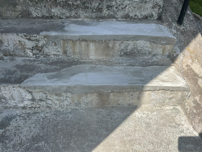 Två trappsteg lagade med betonplåster, ytan ser flammig och ojämn ut, det finns skuggor och synliga sprickor i den omgivande betongen.