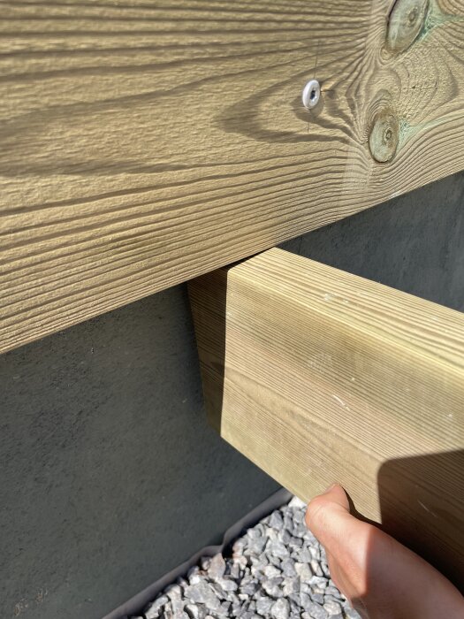 Träregel fäst med skruv mot en betongplatta, med en hand synlig som håller i stödbenet, ovanpå grus.