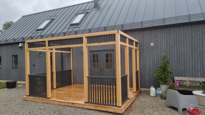 Träveranda med metallräcke monterat mot en grå byggnad, omgivande stenbelagd gård och trädgårdsdekorationer.