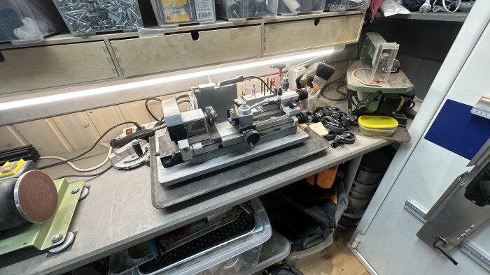 Arbetsbänk med en Unimat 3 metallsvarv och verktyg. Förvaringslådor syns under bänken. Omgiven av verktyg, smärgel och tillbehör.
