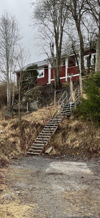 Utomhustrappa i metall och trä leder uppför en sluttning mot ett rött hus. Trappan saknar kryssförstärkning och verkar behöva renoveras.