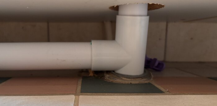 Vit avloppsrör med en L-formad böj genom ett hål i golvplattor, omgiven av lila plastklämmor i bakgrunden.