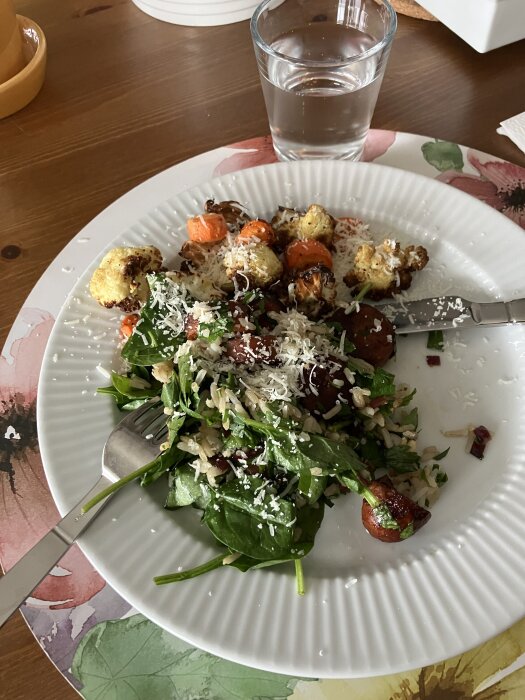 En tallrik med ris, spenat, chorizo, rödlök, toppad med parmesan, bredvid rostad blomkål och morot. Glas med vatten i bakgrunden.