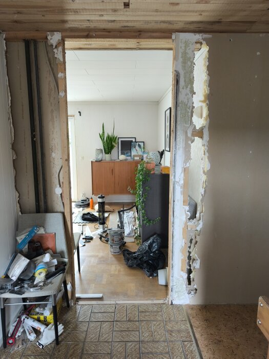 Riven innervägg mellan rum, synliga reglar och verktyg på golvet; hylla och växter i bakgrunden. Renoveringskaos med diverse material.