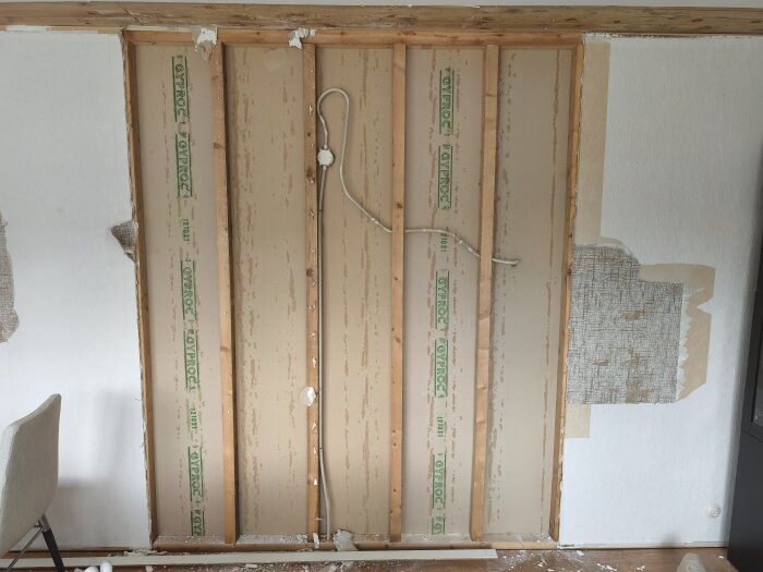 Vägg delvis riven, visar träreglar och gyproc-plattor. Synlig kabel och kvarvarande tapetrester på den borttagna gipsväggen.