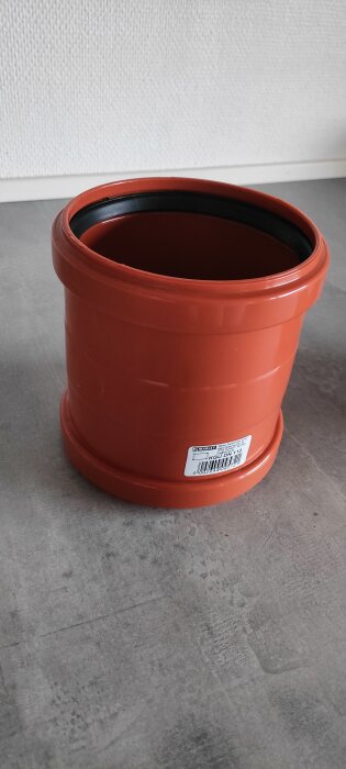 Röd plaströrkoppling med en etikett, placerad på ett grått underlag.