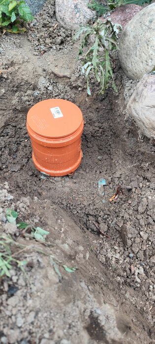 Orange plastbehållare i en grävd grop med sten och jord, omgiven av några växter och löst grus.