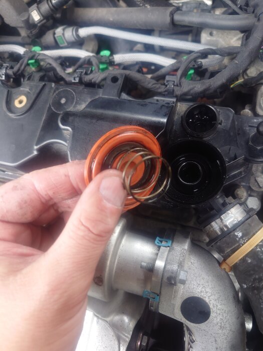 En hand håller ett orange membran med fjäder framför en motor, fokus på detaljer i motorrummet.