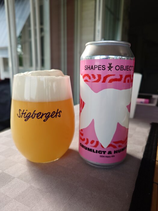 Ölglas med skummande öl bredvid en burk dekorerad med texten "Shapes & Objects" och "Grumligt & Humligt" på ett bord.