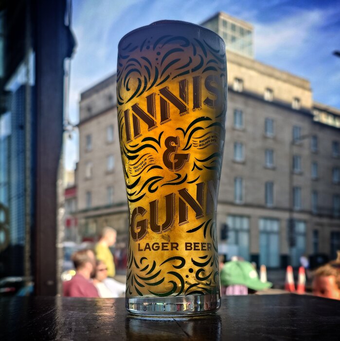 Ett glas Innis & Gunn lager på ett bord utomhus, bakgrund av byggnader och klar blå himmel.