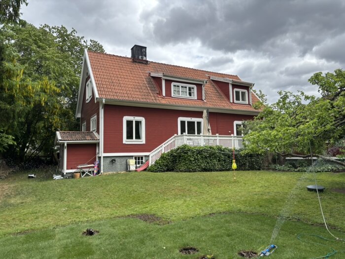 Rött hus med vit veranda och tegeltak, omgiven av grön gräsmatta och träd. Himlen är molnig och föremål står på verandan.