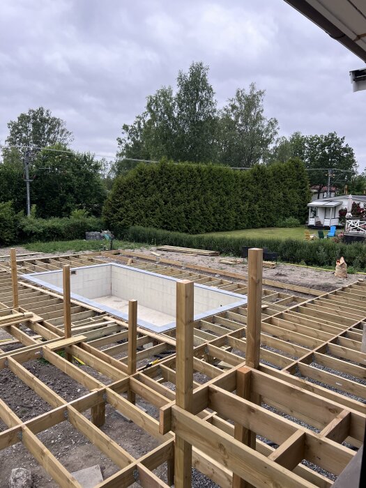 Träramverk för en altan under konstruktion, med reglar runt en nedgrävd trappa eller pool, omgiven av grönska och träd.