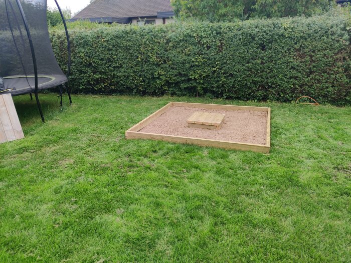 Nybyggd sandlåda med träram fyllt med sand och litet bord i mitten, omgivna av gräsmatta och buskage i bakgrunden.