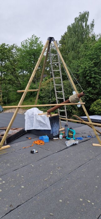 Ställning på tak med stege och båtwinsch för att lyfta ett rostigt stålrör, omgivet av verktyg och material.