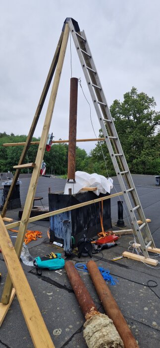 Stålskorsten monterad på taket med hjälp av en båtvinsch och stege, omgiven av verktyg och avskurna rördelar.