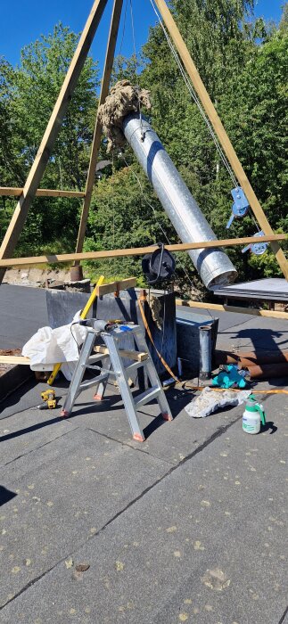 Metallrör upphissad med båtwinsch på ställning, verktyg och material spridda på taket, med gröna träd i bakgrunden.